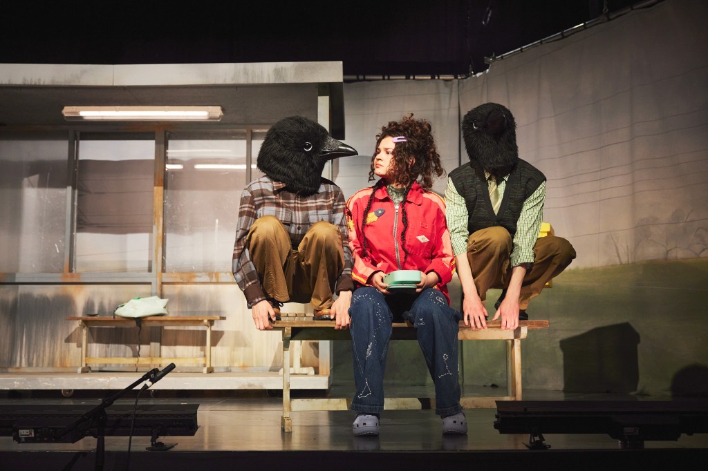 „Zwei Schauspieler stehen auf der Bühne, umgeben von Krähenfiguren; eine Szene aus ‚Kri‘.“